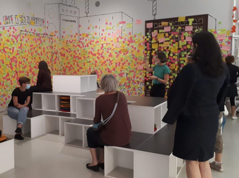 Workshop-Sequenz im hdgö mit Besucher*innen auf weißen Blöcken sitzend und bunten Post-Its auf den Wänden