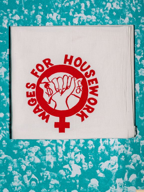 Ein weißes Handtuch auf blauem Grund mit dem roten Aufdruck "Wags For Housework" aus der Arbeitswelt Ausstellung DASA