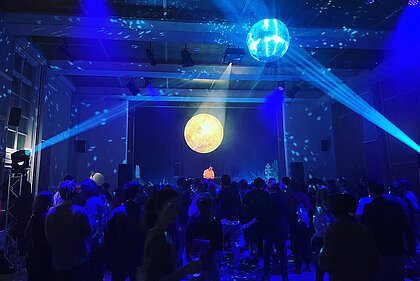 A musician is standing on the stage in the Heimatsaal in the Folk Life Museum, people are standing in front of the stage. The room is in party light.