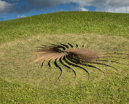 Sole d'acciaio, "sun of steel", is what the artist calls this sculpture. It is an arrangement of 30 curved steel rays around a flint dome. The resulting shapes look like heavy petals in a warm rust red, which change their appearance depending on the incidence of light.