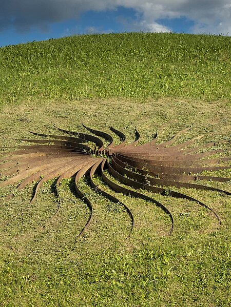 Sole d'acciaio, „Sonne aus Stahl“, nennt der Künstler diese Skulptur. Sie ist eine Anordnung von 30 geschwungenen Stahlstrahlen rund um eine Feuersteinkuppel. Die sich ergebenden Formen wirken wie schwere Blütenblätter in warmem Rostrot, die je nach Lichteinfall ihre Erscheinung ändern.