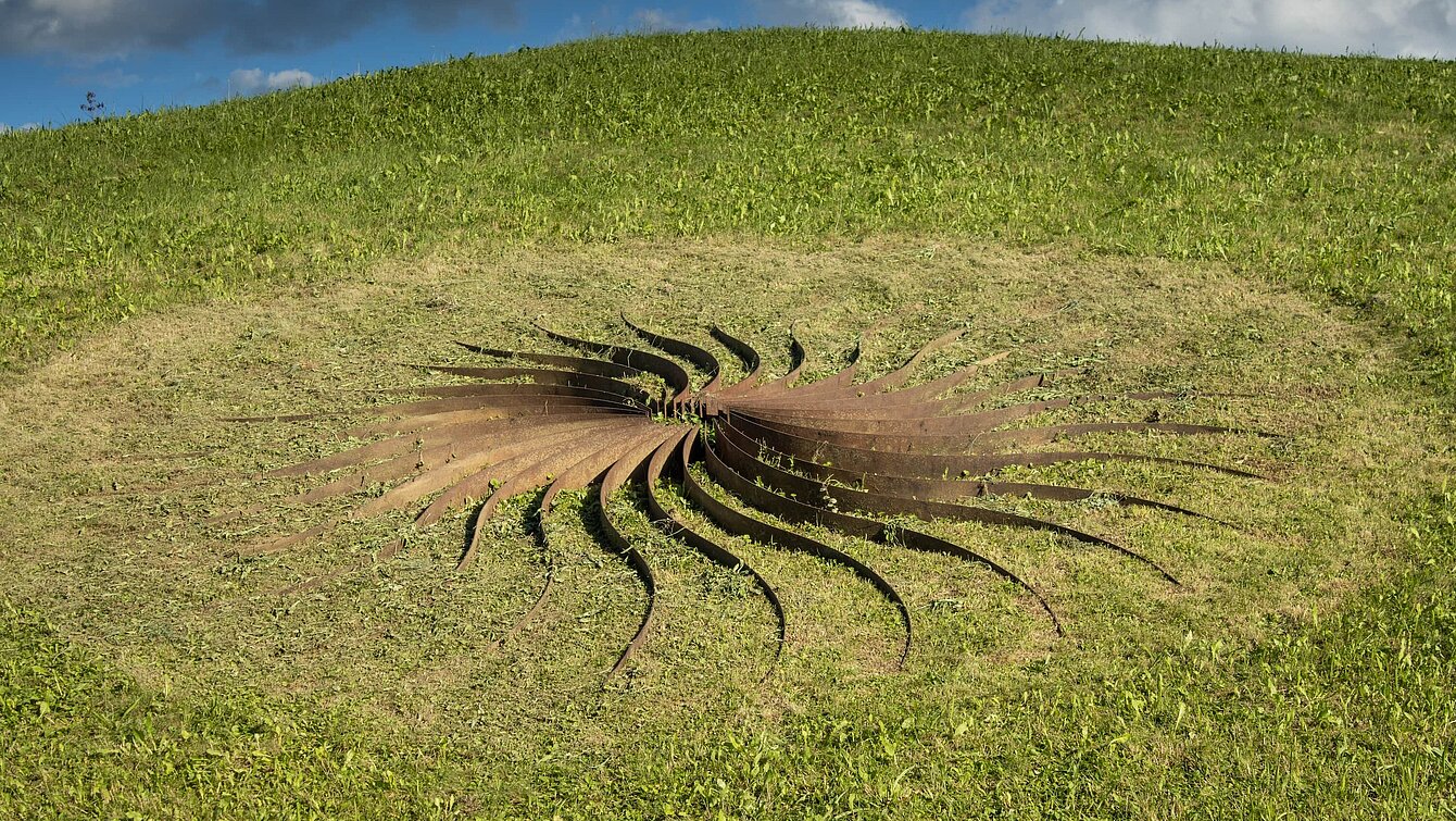 Sole d'acciaio, "sun of steel", is what the artist calls this sculpture. It is an arrangement of 30 curved steel rays around a flint dome. The resulting shapes look like heavy petals in a warm rust red, which change their appearance depending on the incidence of light.