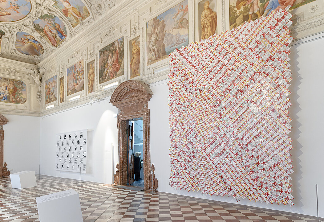 Foto von dem Marmorsaal in Schloss Trautenfels. An der Decke sind bunte Fresken zu sehen. An der Wand hängt eine sehr große Kunstinstallation. Es handelt sich um ein großes Quadrat, das aus vielen kleinen roten und gelben Formen zusammen gesetzt ist.