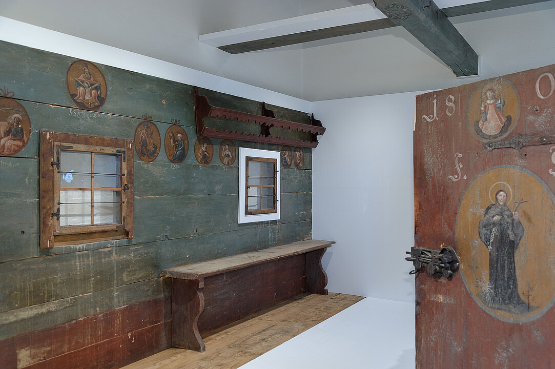 Ausstellungsansicht von einem weißen Raum in dem eine Holzwand aus einem ehemaligen alten Gebäude und eine Tür ausgestellt sind. Die Wand ist blau und rot und mit kleinen Heiligen-Darstellungen bemalt.