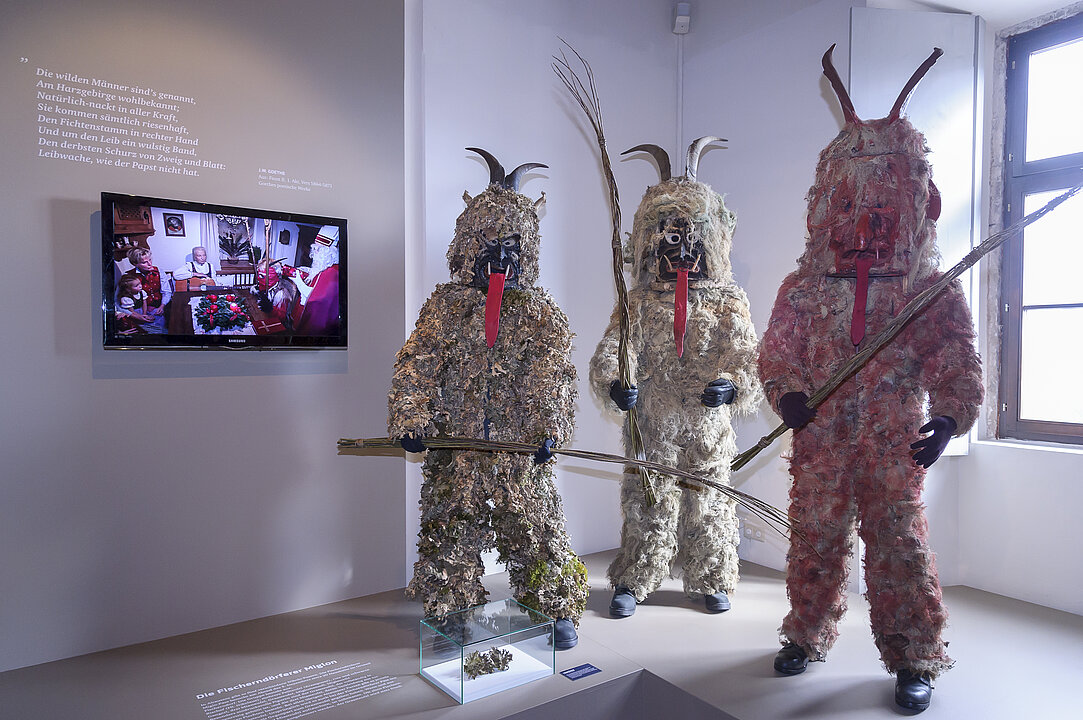 Ausstellungsansicht von einem Raum in dem drei Faschingsfiguren (Miasteufel und Grassteufel) stehen. Daneben ist ein Bildschirm an der Wand angebracht.