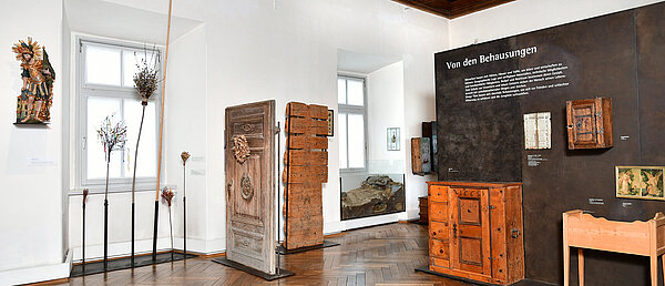 Photography of an exhibition room. In front of a white and a brown wall are various objects e.g. on the left a statue of St. Florian, various palm bouquets, a house door.