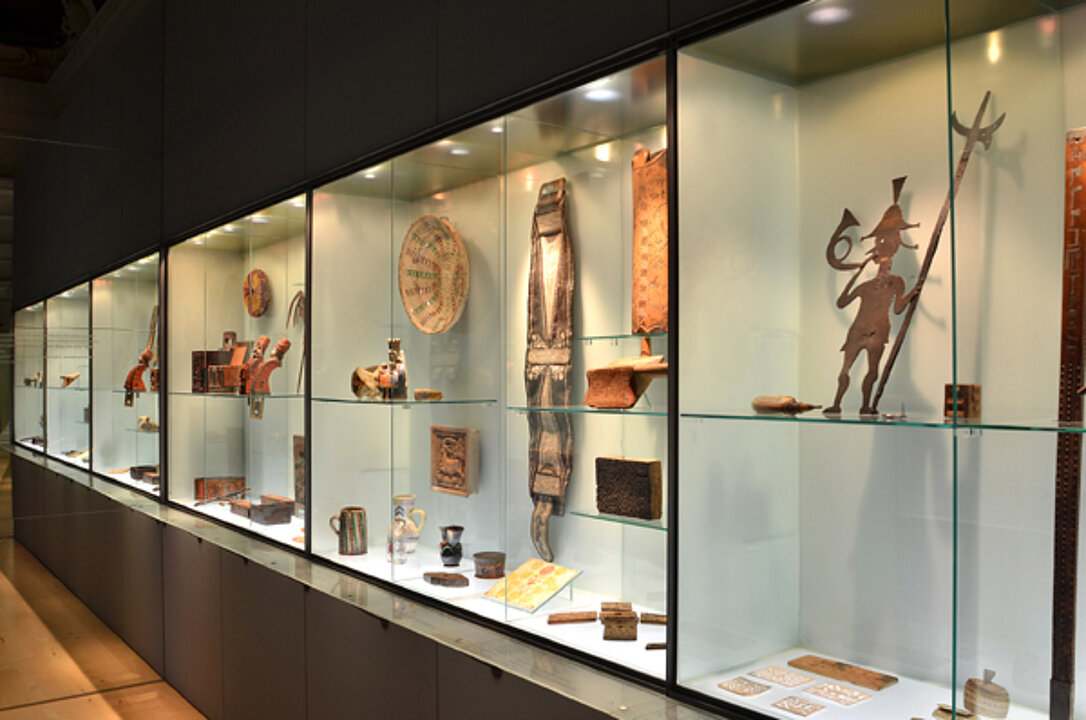 Photo of a long glass display case. In it lie various objects. These all have a use e.g. a bowl or a jug, but are all also specially decorated.