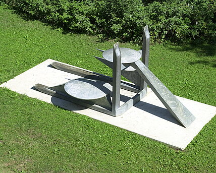 The center of the sculpture is formed by the two vertical and horizontal beams on which the construction rests. The forms appear to be in a finely balanced equilibrium and radiate a certain lightness despite the heaviness of the material. The work gains its concrete meaning by comparing the individual components of the sculpture with generally known objects.