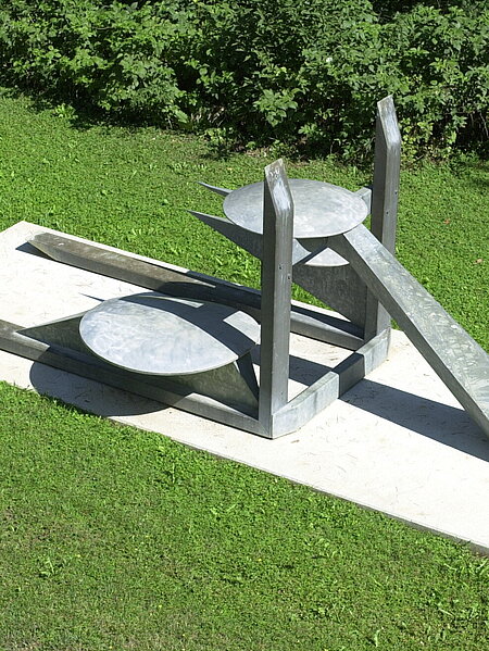 The center of the sculpture is formed by the two vertical and horizontal beams on which the construction rests. The forms appear to be in a finely balanced equilibrium and radiate a certain lightness despite the heaviness of the material. The work gains its concrete meaning by comparing the individual components of the sculpture with generally known objects.