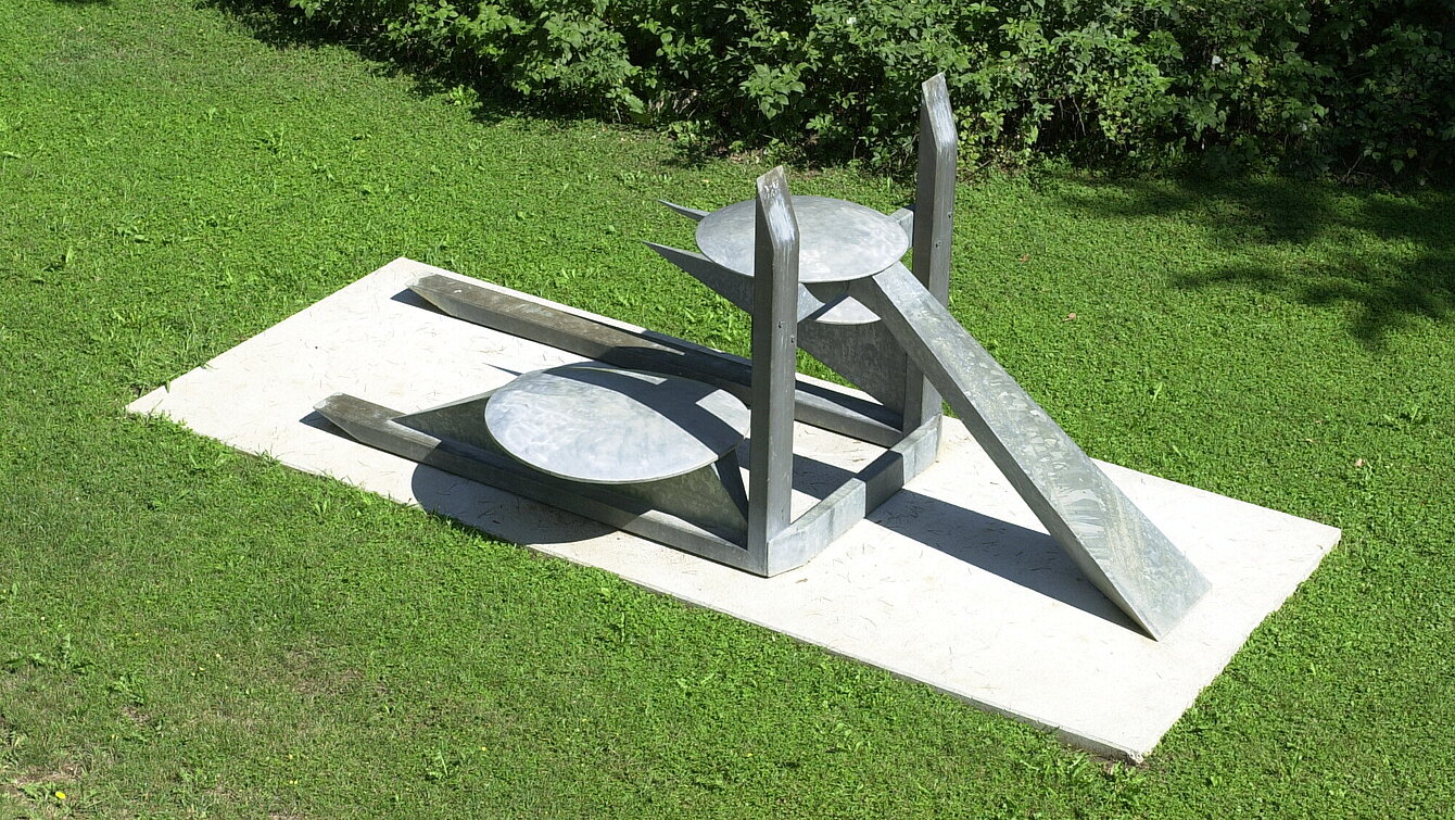 The center of the sculpture is formed by the two vertical and horizontal beams on which the construction rests. The forms appear to be in a finely balanced equilibrium and radiate a certain lightness despite the heaviness of the material. The work gains its concrete meaning by comparing the individual components of the sculpture with generally known objects.