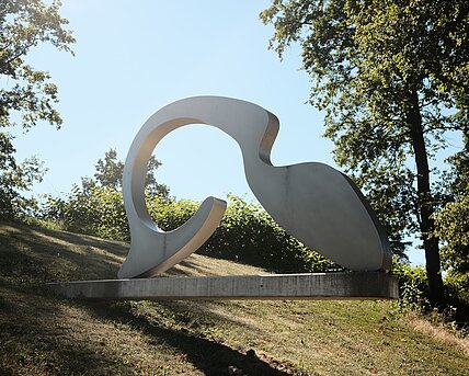 Ursprünglich computergenerierte Skulptur, die aus Metall gefertigt wurde. Sie abgerundet geformt und liegt auf einem Sockel aus Beton, der aus einem abfallenden Hang hervorsteht.