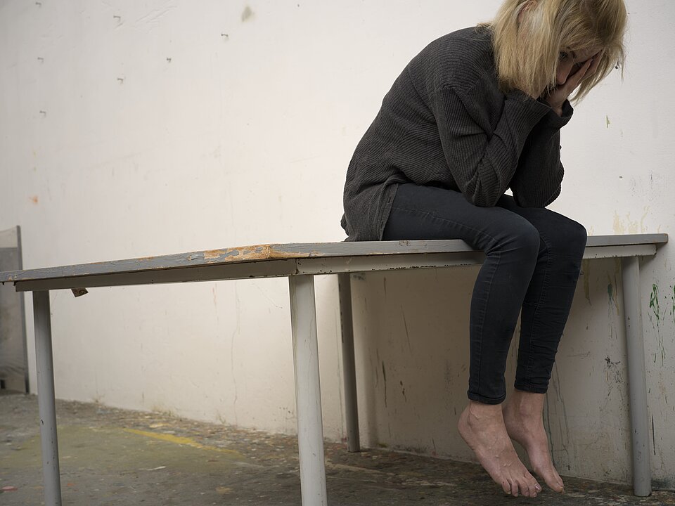 Auf dem Foto sieht man eine Frau auf einem Tisch sitzen. Die Frau mit blonden kurzen Haaren ist schwarz gekleidet und hat die Hände ins Gesicht geschlagen.