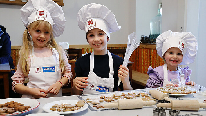 Drei Kinder mit weißen Bäckerhüten und Schürzen sitzen am Tisch und backen gemeinsam Kekse; vor ihnen liegen Teig, Ausstechformen und fertige Kekse.
