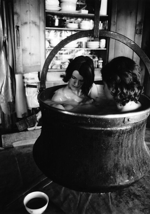 Zwei junge Frauen, sitzen in einem sehr großen Topf, der von der Decke hängt. Nur ihre Köpfe und Schultern schauen aus dem Wasser heraus.