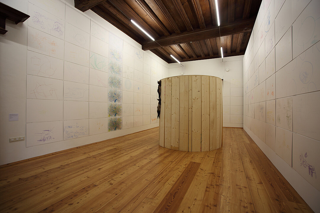 Ausstellungsansicht von einem langen schmalen Raum. Die Wände sind von oben bis unten mit weißen Platten bedeckt auf denen bunte Zeichnung sind, die Kinderzeichnungen ähneln. In der Mitte des Raums steht eine ca. zwei Meter hohe runde Wand aus hellem Holz.
