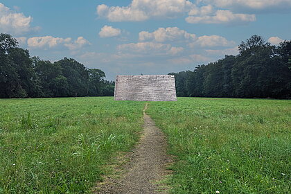 Grüne Wiese in der unteren Hälfe des Fotos mit einem Weg in der Mitte hin zu einem Flachszelt. In der oberen Hälfte sind am Wiesenrand Bäume im Hintergrund zu sehen wie darüber der blaue Himmel mit Wolken