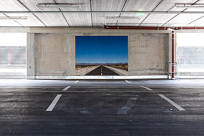 Auf der kargen Betonwand – wie in einer Unterführung – leuchtet ein Foto: eine einsame Straße, die sich durch eine weite Steppe windet, über ihr ein strahlend blauer Himmel.