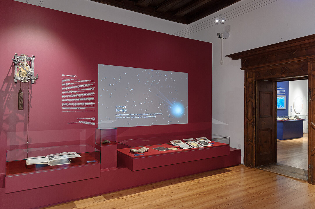 Ausstellungsansicht von einem Raum mit einer roten Wand. Auf die Wand wird eine Video projiziert, das einen Meteorit in der Galaxie zeigt. 