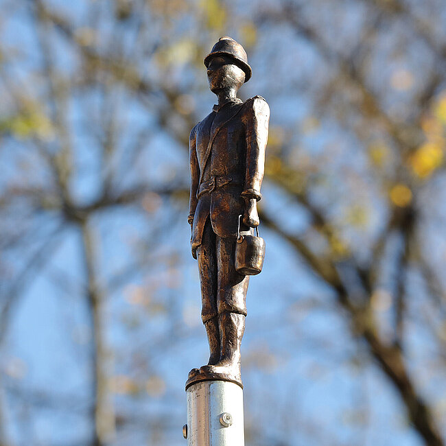 Ein Feuerwehrmann als Bronzeguss. Im Hintergrund Äste vor einem blauen Himmel.