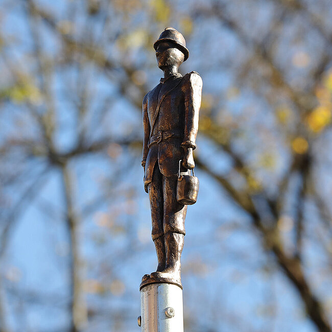 Ein Feuerwehrmann als Bronzeguss. Im Hintergrund Äste vor einem blauen Himmel.