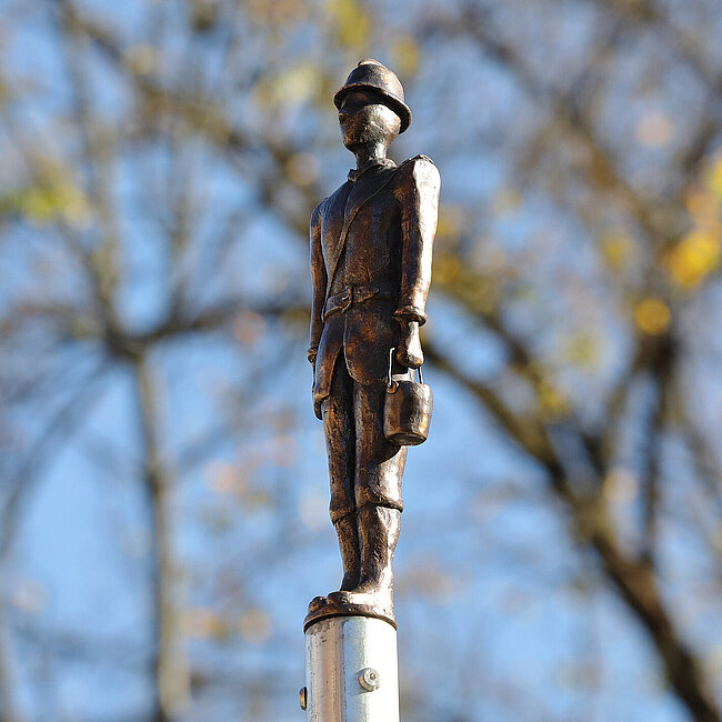 Ein Feuerwehrmann als Bronzeguss. Im Hintergrund Äste vor einem blauen Himmel.
