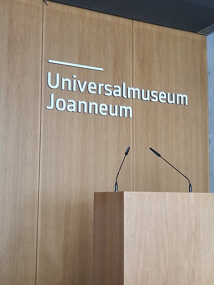 Das Rednerpult im Auditorium um Joanneumsviertel steht auf dem Bühnenpodest mit dem Schriftzug Universalmuseum Joanneum auf Rückwand.