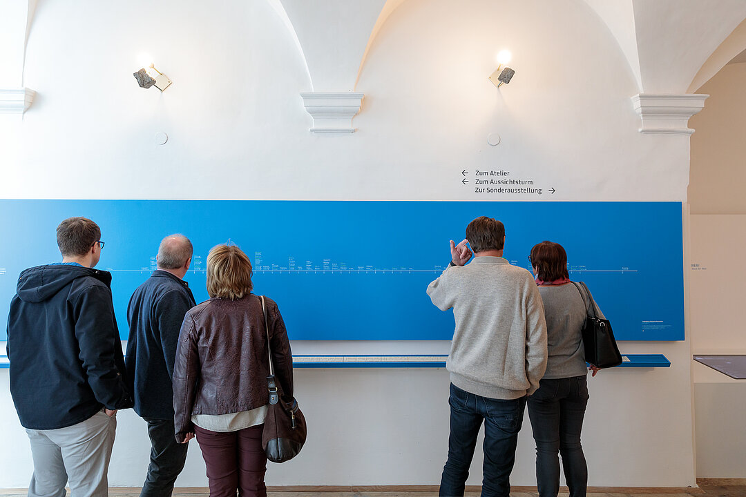 Foto von der Ausstellungseröffnung. Menschen stehen in einem Raum und lesen Texte an den Wänden.