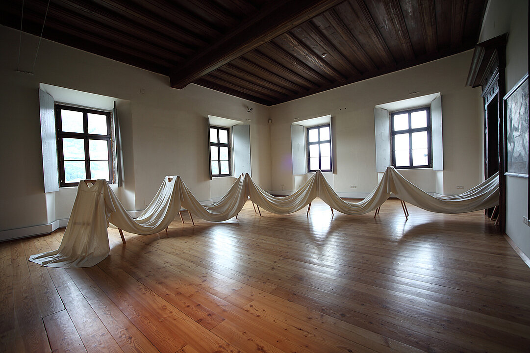 Ausstellungsansicht von einem Raum mit vier Fenstern. In der Mitte des Raums stehen sechs Gestelle über denen ein langes weißes Tuch liegt.