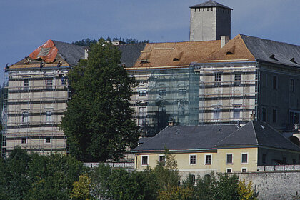 
Photography of Trautenfels Castle. There is scaffolding on the castle walls. There are craftsmen on the roof re-doing the roof. Parts of the roof are already light brown with new shingles, parts are still dark brown with old shingles.