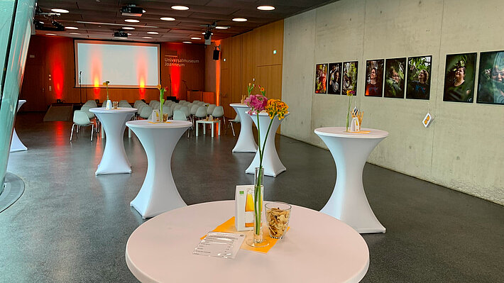 Möbelaufbau für einen Empfang mit Vortrag im Kurt Jungwirth Auditorium. Im vorderen Bereich stehen Stehtische mit Dekoration. Im hinteren Bereich ist eine Reihenbestuhlung vor dem Podium. An den Wänden hängen Bilder und das Auditorium ist in stimmungsvolles Licht getaucht.