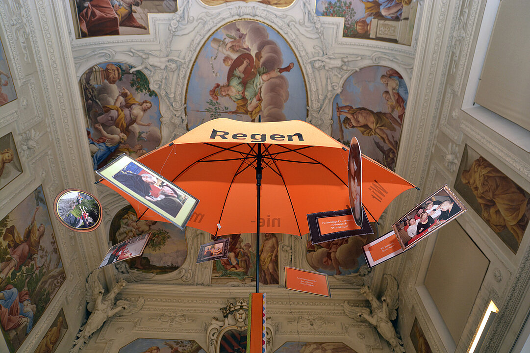 In einem Raum hängt ein orangener Regenschirm von der Decke. Die Decke ist mit Malereien und Stuck verziert.