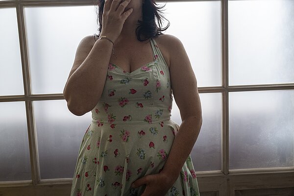 Ein Foto om Querformat auf dem eine Frau, dunkles schulterlanges Haar mit einem weißen ärmellosen Kleid abgebildet ist. Die Frau hält die rechte Hand vor ihren Mund. Das Gesicht der Frau ist nicht vollständig zu sehen.  