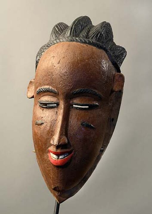 Eine Maske von einem länglichen Schwarzen Kopf mit schmalen Augen, schmaler Nase und einem roten Lippen.