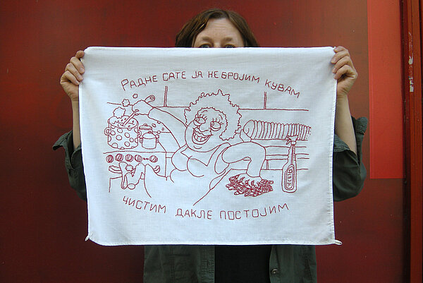 Eine Frau vor rotem Hintergrund hält ein besticktes Tuch vor ihr Gesicht, in dem eine Karikatur einer Frau in einer Küche zu sehen ist, die gleichzeitig kocht und putzt. Ergänzt wird dieses Bild durch einen kyrillischen Titel.