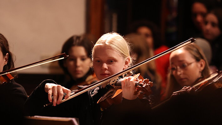 Eine junge Violinistin spielt konzentriert ihr Instrument, umgeben von weiteren Orchestermitgliedern im Hintergrund. Der Fokus liegt auf ihr und ihrer Geige, während die übrigen Musiker*innen leicht unscharf erscheinen.