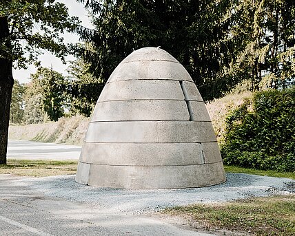 Ein Betonbunker in Form eines Bienenkorbs oder Sprengkopfes mit drei senkrechten Öffnungen. Diese ähneln Schießscharten. Oben befindet sich eine Luke aus Metall.