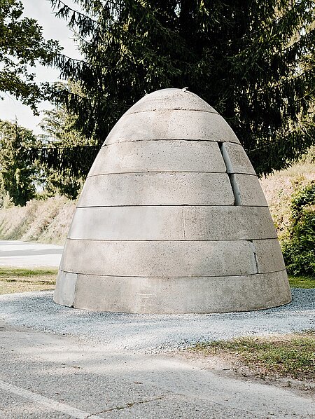 A concrete bunker in the shape of a beehive or warhead with three vertical openings. These resemble embrasures. There is a metal hatch at the top.