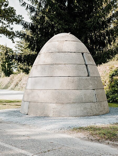 Ein Betonbunker in Form eines Bienenkorbs oder Sprengkopfes mit drei senkrechten Öffnungen. Diese ähneln Schießscharten. Oben befindet sich eine Luke aus Metall.