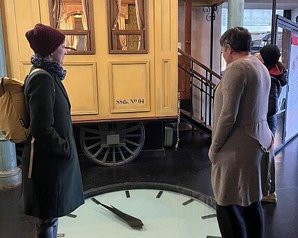 2 Personen sind in Rückenansicht gezeigt, am Boden befindet sich eine Uhr.