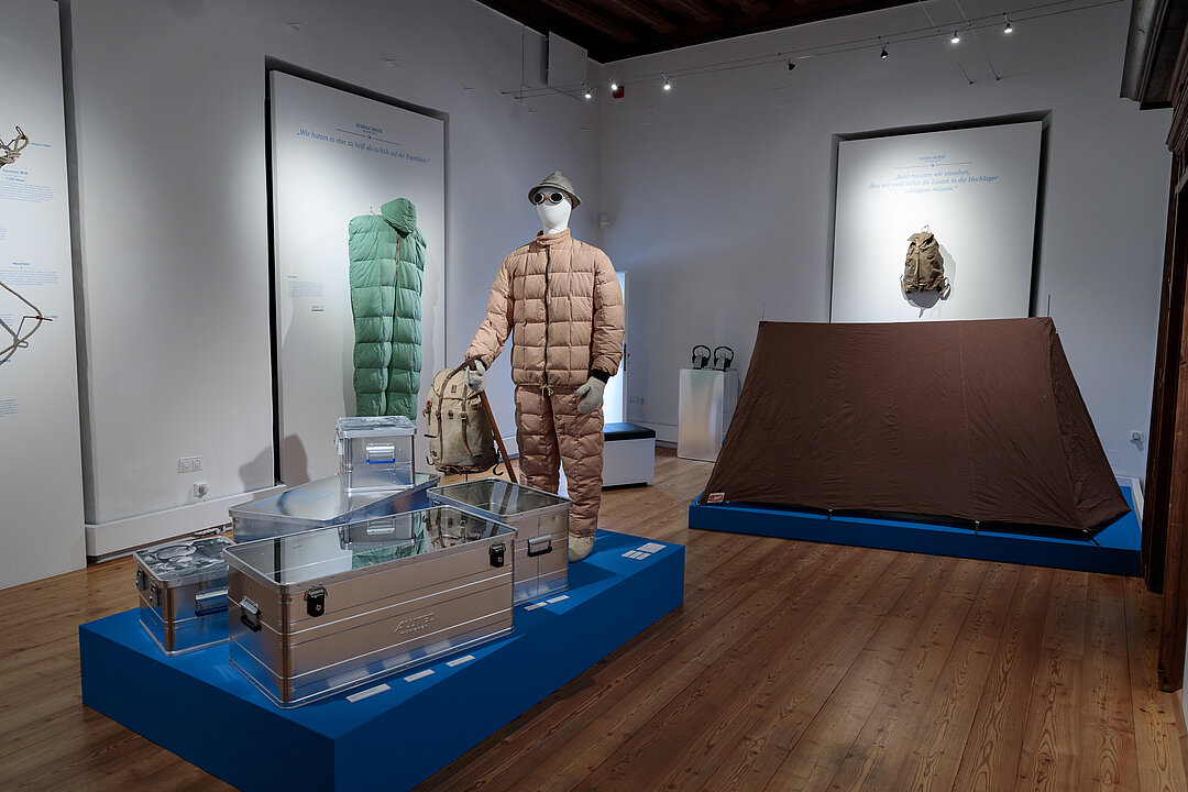 Ausstellungsansicht: in der Mitte des Raums steht ein Podest mit silbernen Boxen. Dahinter steht eine Schaufensterpuppe mit beigem Bergsteiger-Outfit.