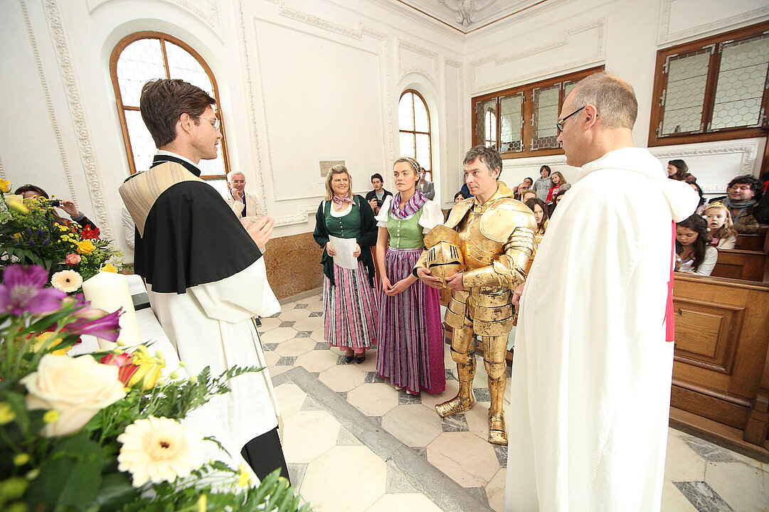 In einer Kirche steht ein Pfarrer. Vor ihm stehen vier Personen. Ein geistlicher im weißen Gewand, ein Mann mit goldener Rüstung und zwei Frauen in Tracht. Dahinter sitzen auf Bänken, das Publikum.