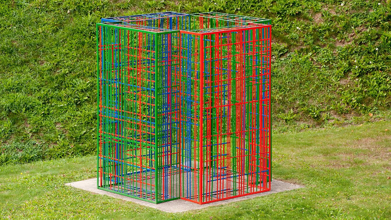 Die Skulptur besteht aus farbigen Metallstreben steht auf einem Sockel. Vier farbigen Module aus Metall überlagern und vernetzen sich je nach Blickwinkel immer anders.