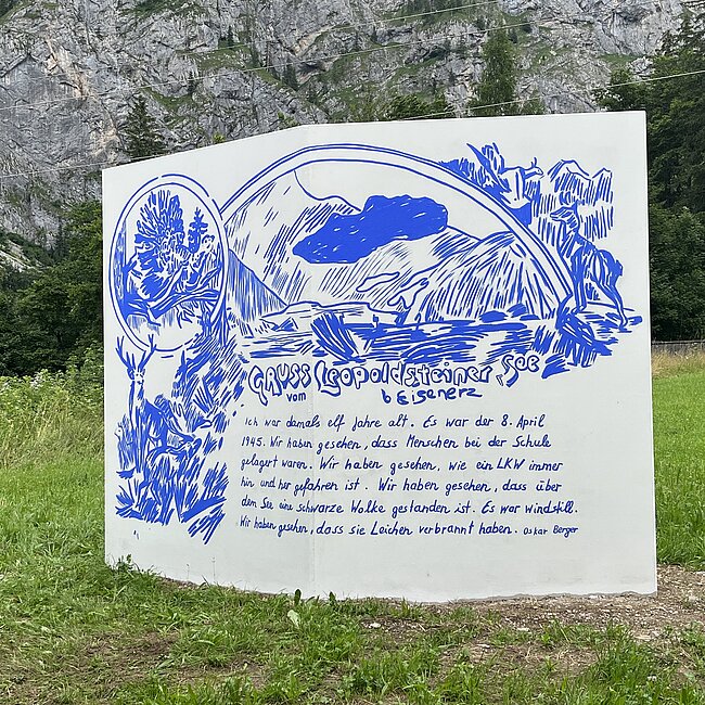 [Translate to English:] Auf einer Wiese steht eine weiße Skulptur wie eine Seite von einem Buch, in der Mitte ist diese rechteckige Wand leicht geknickt. Darauf ist in Blau eine Zeichung mit den Worten: Gruss vom Leopoldsteiner See bei Eisenerz. Ich war damals elf Jahre alt. Es war der 8. April 1945. Wir haben gesehen, dass Menschen bei der Schule gelagert waren. Wir haben gesehen, wie ein LKW immer hin und her gefahren ist. wir haben gesehen, dass über dem See eine schwarze Wolke gestanden ist. Es war windstill. Wir haben gesehen, dass sie Leichen verbrannt haben. Oskar Berger