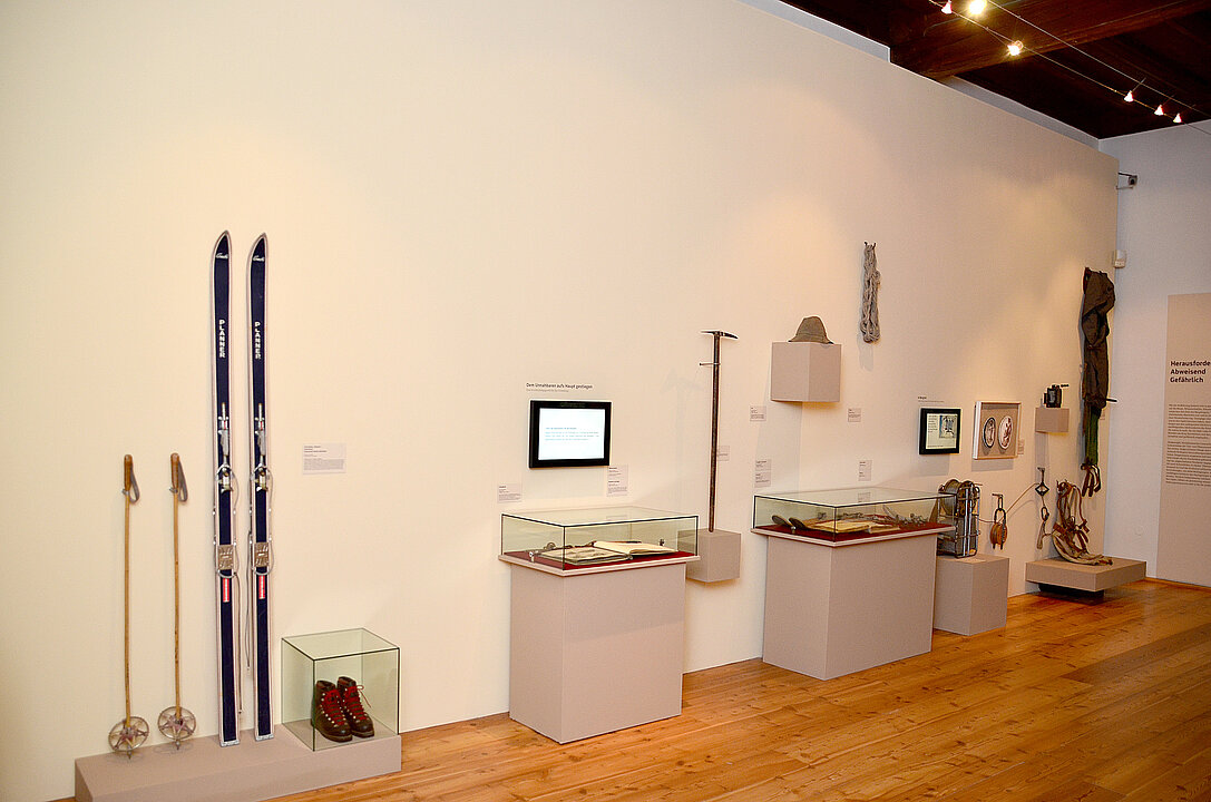 Zusehen ist ein Raum an der Wand stehen auf Sockel und Podesten verschiedene Museumsobjekte z.B. Skier und Stöcke.
