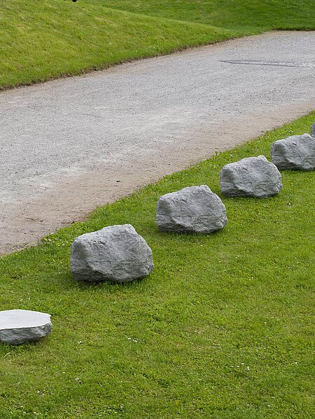 Sechs Steine aus Beton in einer Reihe, die dem ursprünglichen Kalkstein nachempfunden sind. Daneben im gleichen Abstand eine 0,4-fache Version eines der Steine.