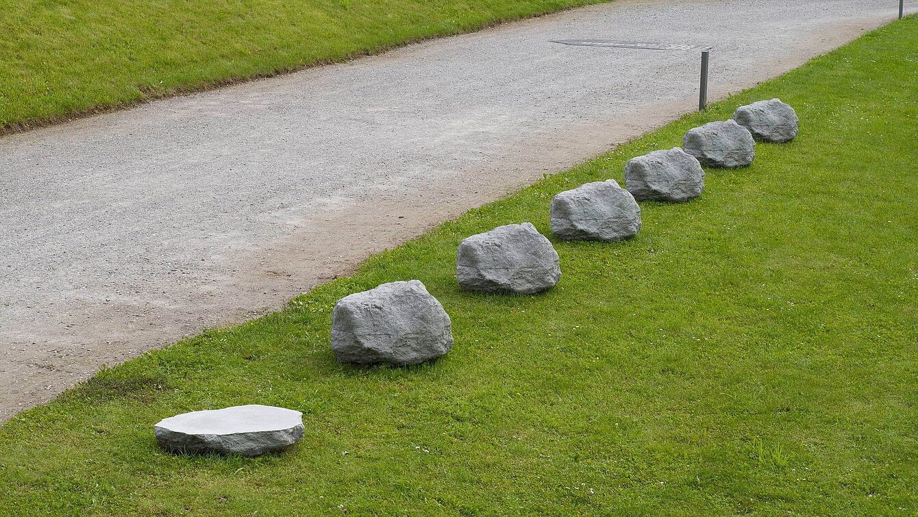 Sechs Steine aus Beton in einer Reihe, die dem ursprünglichen Kalkstein nachempfunden sind. Daneben im gleichen Abstand eine 0,4-fache Version eines der Steine.