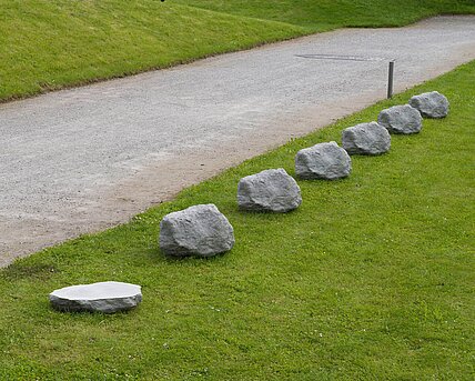 Sechs Steine aus Beton in einer Reihe, die dem ursprünglichen Kalkstein nachempfunden sind. Daneben im gleichen Abstand eine 0,4-fache Version eines der Steine.