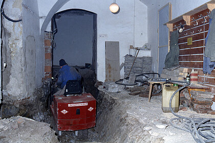 Fotografie von einer Baustelle. Auf der rechten Seite liegen Geräte und Schläuche. In der Mitte sitz ein Mann auf einem roten Fahrzeug in einem Graben.