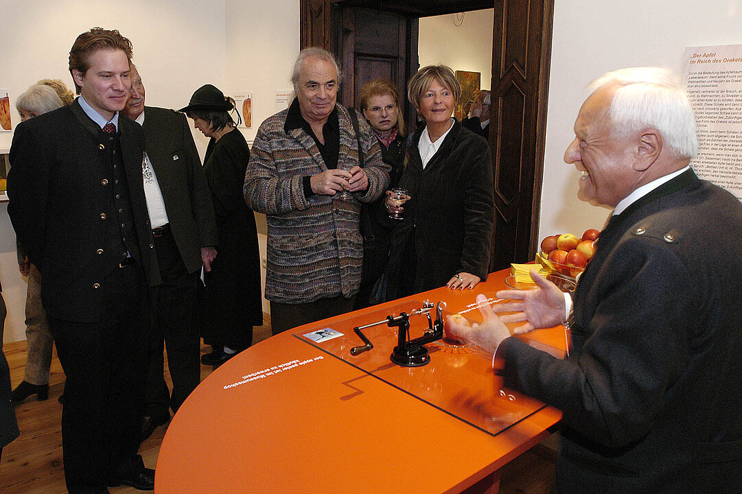 Eine Menschengruppe steht vor einem orangenen Tisch. Davor steht ein Mann und erklärt eine Apfelschälmaschine.