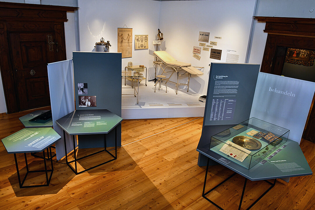 Photo of an exhibition room. In a white corner are objects, for example, a couch from an old doctor's office.